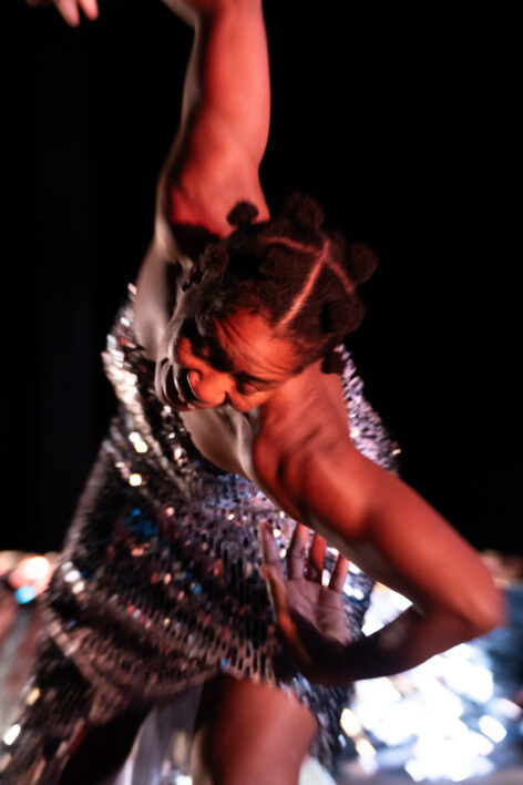 Black dancer in a shimmery strapless silver dress leaning headfirst into the frame, bent at the waist, with arms outstretched and bent at the elbows. Hair is styled in afro puffs and face is animated, eyes looking down and mouth open. Light is reflecting off the dress, brown skin and a shimmery object in the background in the bottom part of the image. The rest of the background is black. Photo by Robbie Sweeny.