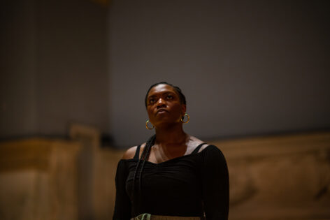 Performance photo of Ogemdi Ude, a dark skinned black woman. She wears her braided hair tied back, gold hoops and an off the shoulder black long sleeve top. She has a calm face expressioN. Her head is tilted up slightly and she looks beyond the camera. Photo by Rachel Keane.