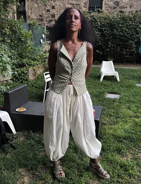 Black women standing with hands behind her back. She is wearing a green and yellow v-neck blouse with white barrel pants. Photo by Orion Gordan.