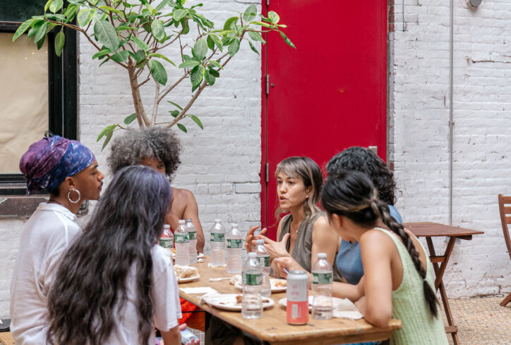 A group of people sit at a table and lean in to speak. In the background a bright red door, Gray brick wall and a small tree. Photo courtesy of the AoCC.