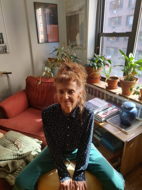 Vicky sits on a big yellow ball in her living room. My grayish brown hair is in a messy pony tail on top of my head. I am wearing a dark blue shirt with pale dots and green sweat pants. In the background, plants, books, a window and a red chair. Photo by Alan Mandell.