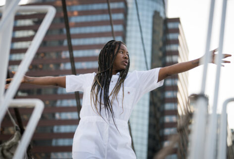A brown skinned performer with long brown braids in a white jumpsuit. The performer's arms are horizontally outstretched and they are looking intently to the left while they stand on the bow of a ship. A high rise building with reflective windows is in the background. Photo by Cherylynn Tsushima.