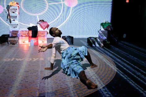 Performance photo of André Zachery dancing in a lunge position, with him wearing a white long-sleeve top and turquoise raffia pants, with aesthetic materials on set on stage, taken by Tony Turner.