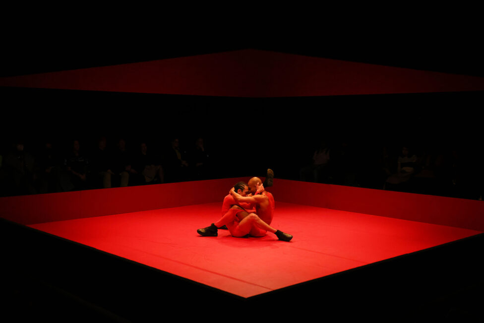  Two performers, bathed in red light, are in a seated embrace. They press their foreheads together, legs intertwined.