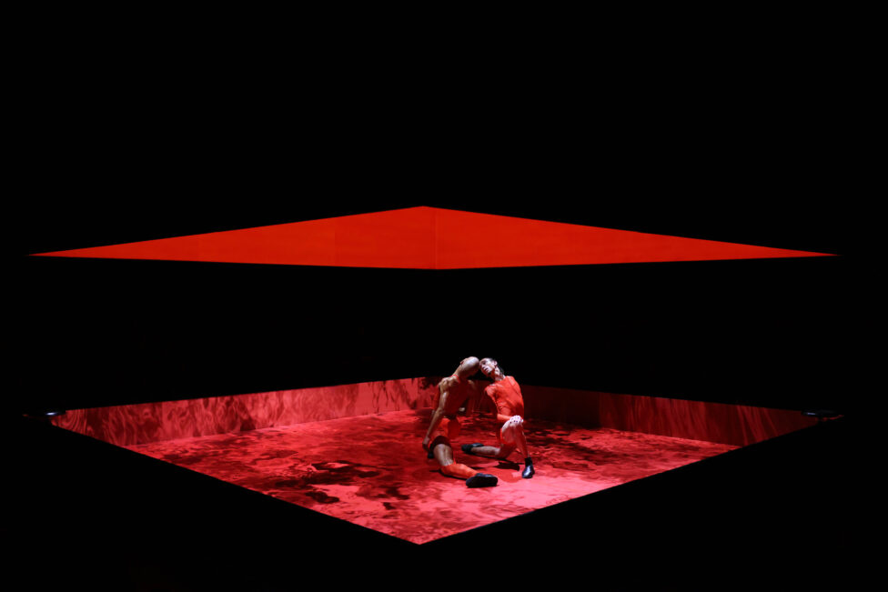 Two dancers in red clothes kneel, leaning in toward each other. Above them on the stage, a vivid panel of red light hangs like a roof.