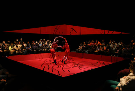 Two men hold hands in the center of a red stage. They are bent away from each others' hands while mirroring each other. The red stage has black markings on it and the audience surrounds the dancers on all sides. There is a large square mirror hanging above the dancer's heads. Photo by Jeremy Lawson, courtesy of MCA Chicago.
