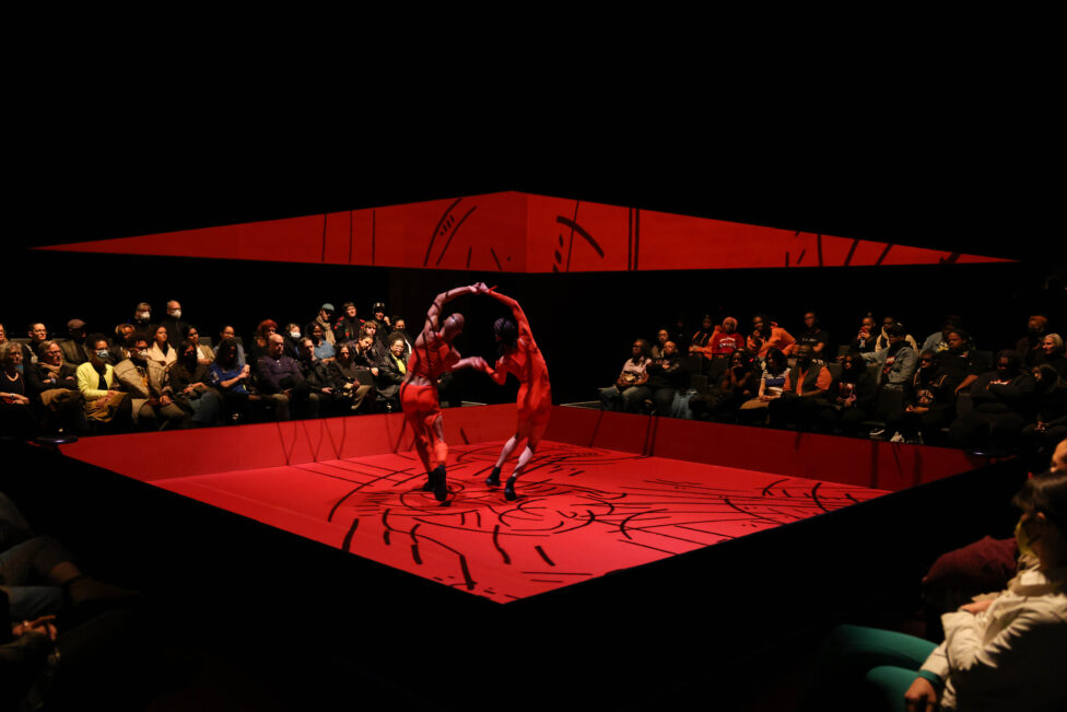Two dancers hold hands, arching toward each other on a red-lit stage. The audience can be seen surrounding the stage in the background. Photo by Jeremy Lawson courtesy, of MCA Chicago