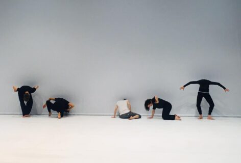 Photo of several dancers posing in front of a gray wall. They pose at different levels, some standing others sitting or on their knees. Photo courtesy of the artist.