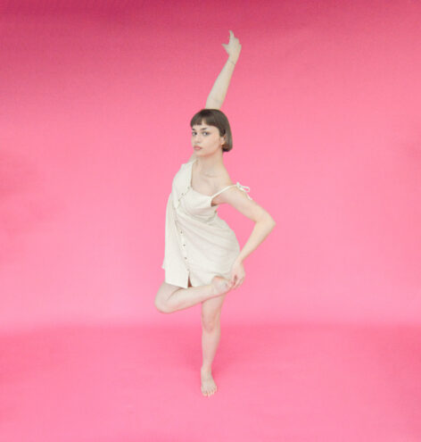 Tatiana wears an off-white sundress as she poses against a hot pink backdrop. She sits in a plié as her left leg is an over-crossed passé with her left arm extended upwards. Her right arm holds her left foot. 
