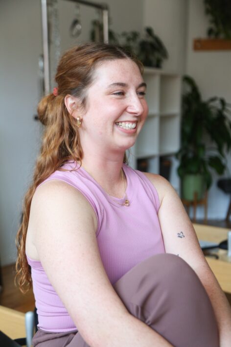 A white woman with long, wavy, red hair is seated looking off to the right. She smiles and squints her eyes. She is inside of a Pilates studio with wood floors and white walls and plants scattered throughout the background. Photo by Rainey Scarborough Courtesy of Greenpoint Pilates Studio