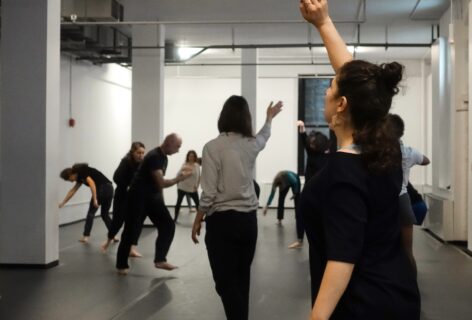 Participants move in various positions with mouths open in sound during a Voice and Movement class in Movement Research's Ninth Street Studio. In the foreground a dancer in black reaches their arm upwards. Photo by Harry Shunyao Zhang.
