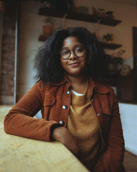 A portrait of Rebecca sitting at a table. She wears black framed glasses, a brown jacket, a tan top, and her hair is down right above her shoulders. Behind her are shelves and plants. Image courtesy of Artist.