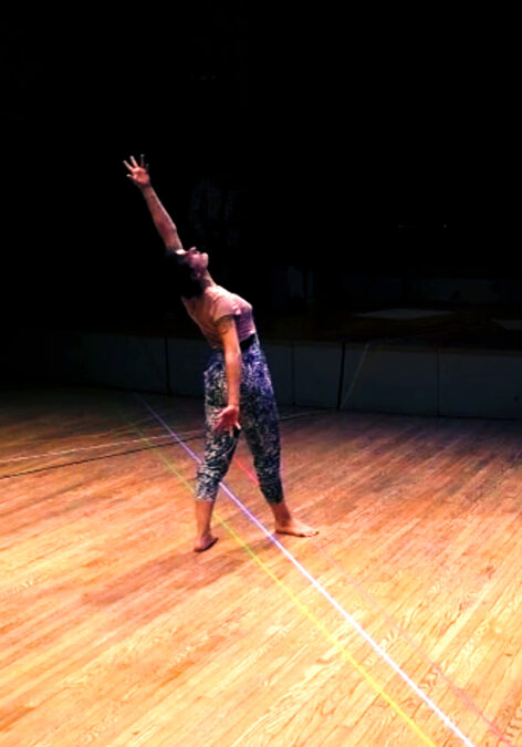 Performance photo of Sarah White-Ayón. A white woman with black hair stands in fourth position with her right foot front and her back facing the audience. She is arching upwards looking towards her left hand that is maximally outstretched above her head while her right hand is outstretched oppositional to her left hand. She stands on a hard wood floor with 3 lines of colored tape, yellow, white and pink attached to the floor passing underneath her feet. She wears black and white speckled pants and a pink t-shirt. The background behind her obscured, almost black. Photo courtesy of the artist.