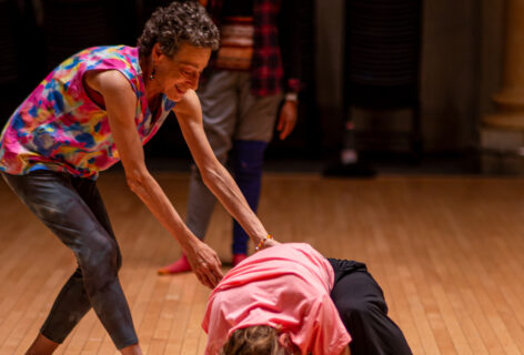 Barbara Mahler guides a student during class. She is extending her arms towards the student while they are crouched on the flood in movement. Barbara is smiling wearing a patterned colorful top. The student wears a bright pink top. Photo by Rachel Keane.