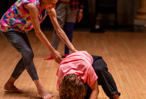 Barbara Mahler guides a student during class. She is extending her arms towards the student while they are crouched on the flood in movement. Barbara is smiling wearing a patterned colorful top. The student wears a bright pink top. Photo by Rachel Keane.