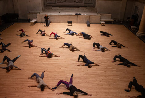 Photo of dancers moving at the Judson Church. Irene Dowd stands int he upper left corner hands on her hips watching the class. The dancers move on the floor creating spirals with their torsos in a unison phrase. Photo by Rachel Keane.