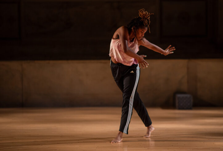 Angie Pittman performing for Movement Research at the Judson Church. She is standing with her legs crossing and her torso twisting to one side. Her arms and gaze look towards her back leg. Photo by Rachel Keane.