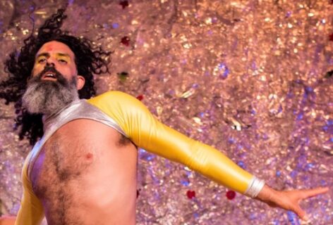 Photo of Antonio Ramos wearing a cropped top with gold sleeves and a silver turtleneck. His chest is bare and his curly hair is flowing freely. He has his mouth slightly opened, arms spread out with yellow and red face makeup. The background is glittery. Photo by Ian Douglas.