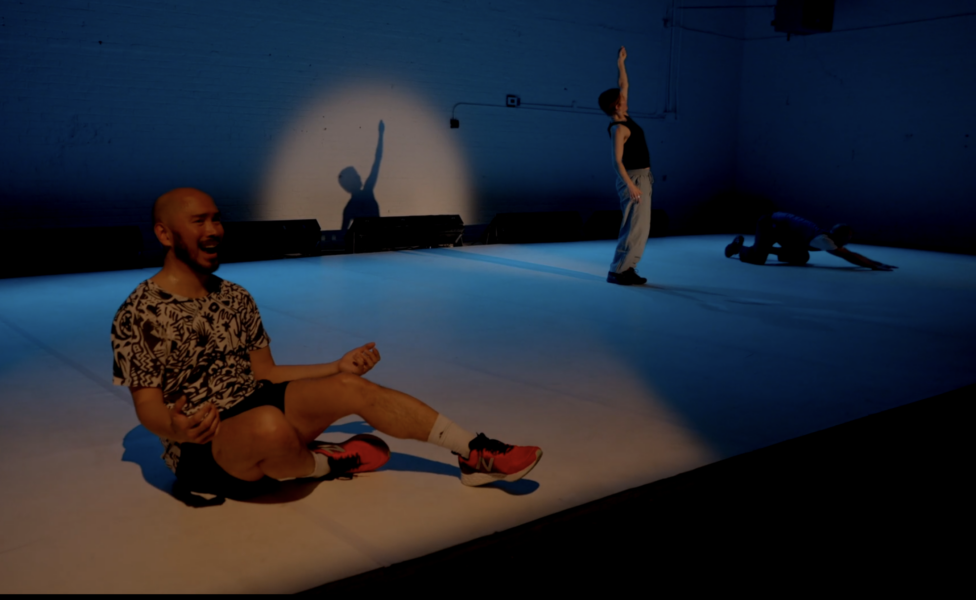 Justin sits in the lit foreground of a darkened, shadowy stage wearing red sneakers, shorts, and a floral button-down shirt. Another dancer stands in a spotlight with left arm reaching upward, creating a shadow behind them. A third dancer crouches close to the ground in the shadows, barely visible. Photo by Peter W. Richards. 