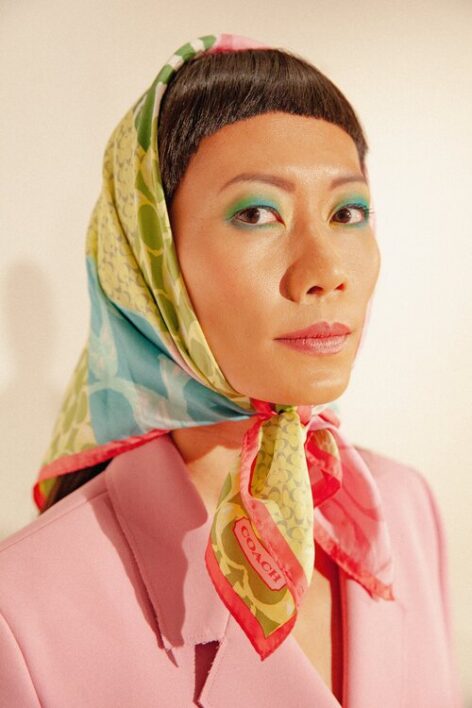 Portrait of Chin-I Chang. She wears a blush pink blazer and a multicolor printed scarf in blue, green and pink around her head and tied under her chin. She has short black bangs and has bright blue eyeshadow on her lids. She looks into the camera with a calm expression. Photo by Eric MacLean.