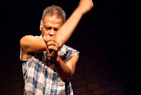 Ishmael Houston-Jones, a brown-skin African-American middle-aged man with short salt and pepper hair is caught mid-fall, crunching up his face and swinging semi-fisted hands in front. He is wearing a tattered blue and white plaid tee-shirt, blue jeans and white sneakers. Photo by Ian Douglas.
