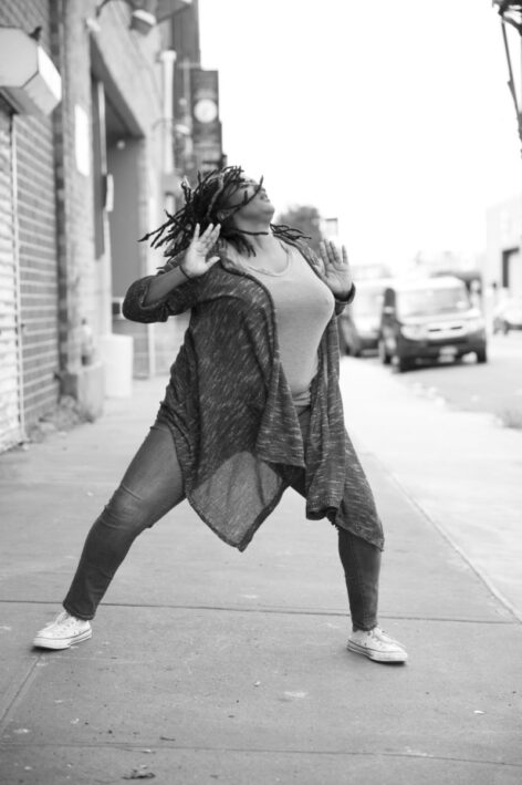 This is a black & white dance image of Kayla Hamilton, who is a dark brown-skinned Black woman. She is throwing her head back as her dreads flow with her as she pushes her arms outward. Her legs are wide and slightly bent. She is wearing jeans and a knee length cardigan that wraps around her thighs. Behind her are storefronts and cars parked on the street. Photo by Travis Magee.