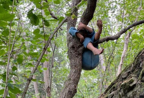 Photo of a person climbing and twisting their body around a tree branch. There is green foliage all around them and they wear a light blue outfit. Photo by Otto Ramstad