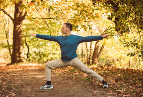 Ori Flomin is in a warrior two position from Yoga practice. He is wearing light beige pants and a blue long sleeve shirt. In this position his right leg is bent to a 90 degree angle while his left leg is fully extended, his spine is vertical and his arms are extended away from each other parallel to the floor. He is standing outside in the park with trees in the background. Photo by Yasmeen Enahora.
