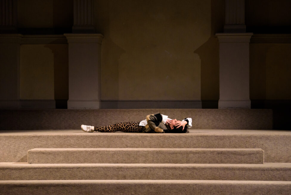 Sam lays on her back at the top of a set of beige stairs, wearing white sneakers, leopard leggings, a white shirt, and a black and gold multi-colored jacket. Her hands touch the top of her head and face. 