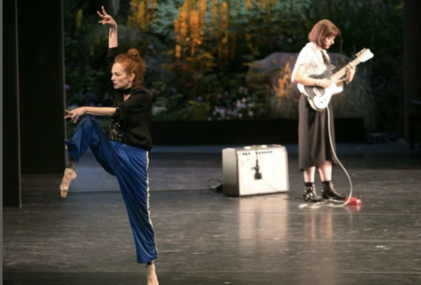 Photo of Jodi Melnick in performance. She balances on one leg, bending her other leg at the knee and raising it towards her torso. One arm is extended in front of her and the other reaches over her head. In the background a musician plays electric guitar with an amp behind them. Photo courtesy of the artist.