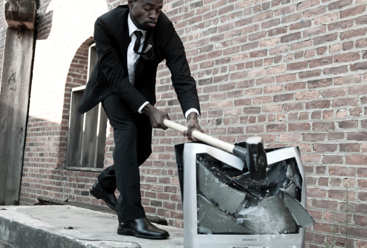André Zachary smashing a television with a mallet. He wears a black suit and tie. In the background, an industrial setting with Brick walls and cement. Photo by Rachel Neville.