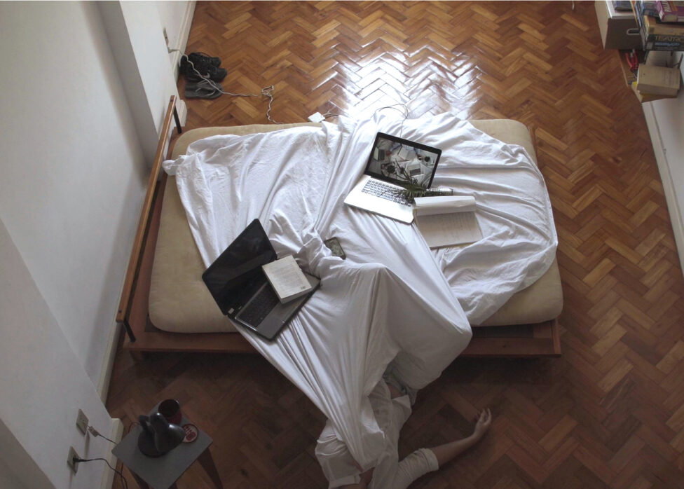 A bed with white sheets and a wooden frame sit in a room with miscellaneous objects around the perimeter. The bottom half of Sofia's body lays halfway on the bed and the top half on the wooden floor. Two laptops and a couple of notebooks remain on the bed's white sheets. Photo courtesy of the artist. 