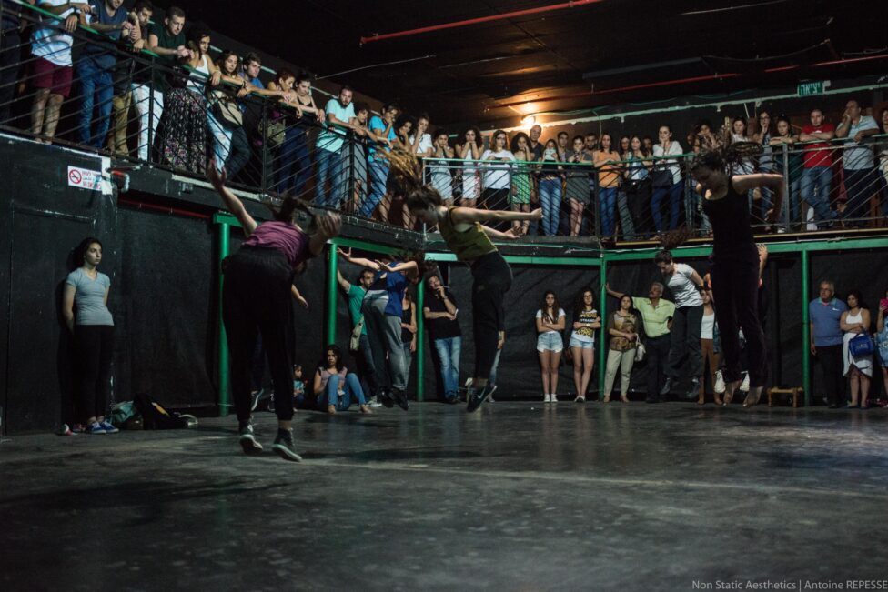 Audience leans over railing and encircles three dancers jumping in a blackbox space. 