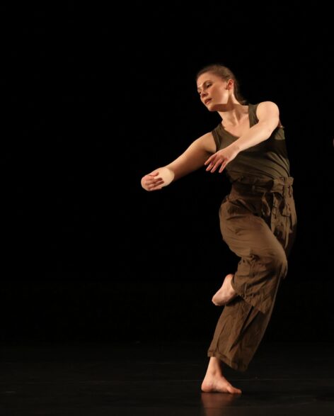 Anna wears a muted, green monochromatic outfit. She is in motion, her body leans from her hips to form a standing c-shaped bend. Her arms extend parallel to the ground and curve together, fingertips reaching together. her upper body twists towards the right, her head and eyes follow the rotation to look just beyond her frame. Photo courtesy of Gamut Dance Company.