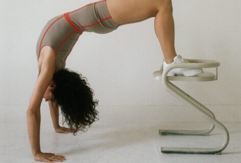 Amelia is white and has brown curly hair. She does a backbend on the ground with her feet on a stool. She has on a short unitard and dance sneakers and is in an empty space. Photo by Jenna Westra.