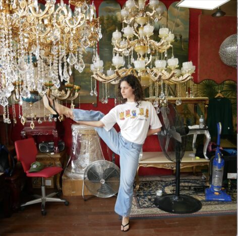 Amelia is white and has brown curly hair. She stands on one leg and holds the other leg with her hand. She is in a chandelier shop. She has on flip flop heels, light wash jeans, and a t-shirt with cowboy shoes on it. Photo by Alexa West.