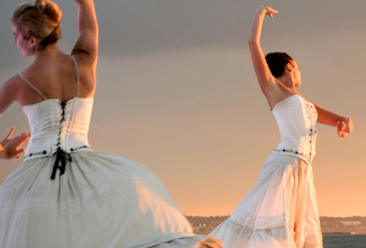 Outdoors at sunset, two barefoot dancers wearing flowing white dresses with dark trim and upswept hair stand at a distance while in similar motion, facing away from each other with heads turned to the side, one arm arched above their heads with a palm facing up and the other bent towards their sides with a palm out. Photo AI generated, courtesy of the artists.