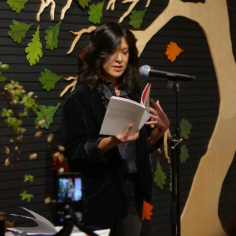 A multi-racial woman with medium-length black hair stands behind a microphone while she reads aloud from a book. She’s dressed in many shades of black and gestures with one hand while she holds a book with the other. There’s a wood carving of a tree losing its leaves in the wind behind her. Photo courtesy of the artist.