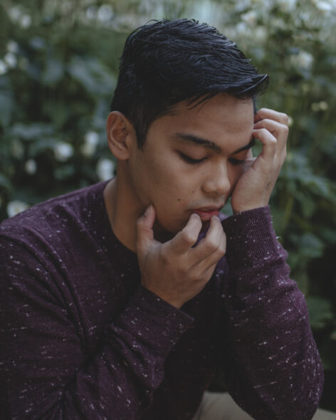 A side profile of Miggy, who crouches amidst bushes and white flowers that recede blurrily into the background. Miggy’s fingers gently crawl up from a long-sleeved maroon shirt, over chin and lips, and toward a small ocean of black, wavy hair. The brown skin of Miggy’s cheek is caressed by the palm of a hand, supporting closed eyes that look down in contemplation. 