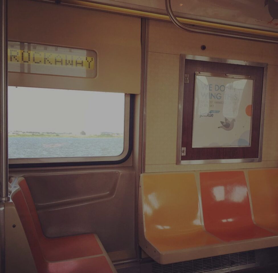 [The interior of a subway cart passing over a body of water with orange and yellow seats; two facing the left and three facing the front. Above the three seats, hangs a poster ad. The screen above the window indicates that the train heading to ‘ROCKAWAY’.]