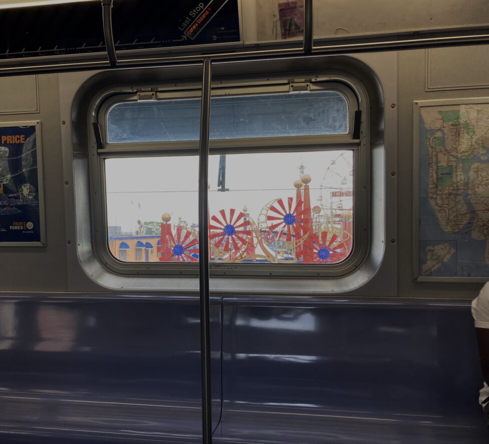 [The interior of a subway cart at its last stop on Coney Island, as indicated on the screen above a window. The window overlooks a colorful amusement park. The bench seating is of a blue color, and a map of New York City hangs to the right of the window.]