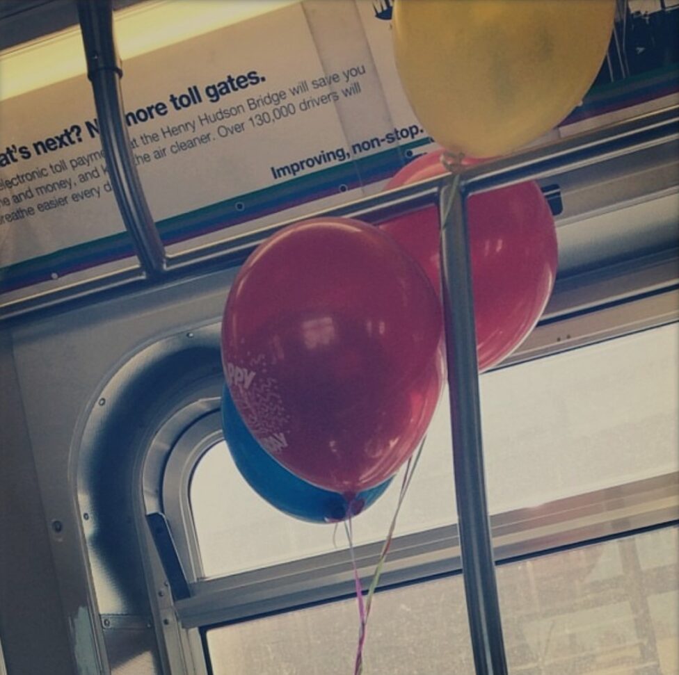 [Two red, one blue, and one yellow colored balloons hover amongst the metal bar of a subway cart.]