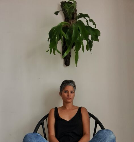 Betina is sitting underneath a plant hung on wall. She is wearing a black tank top and jeans. 