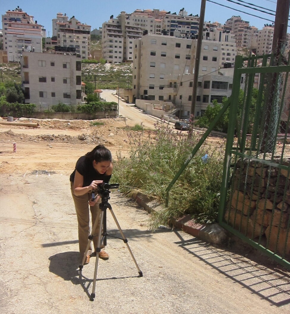 Samar leans forward over a tripod, sand-colored 8-10 story buildings loom in the background. 