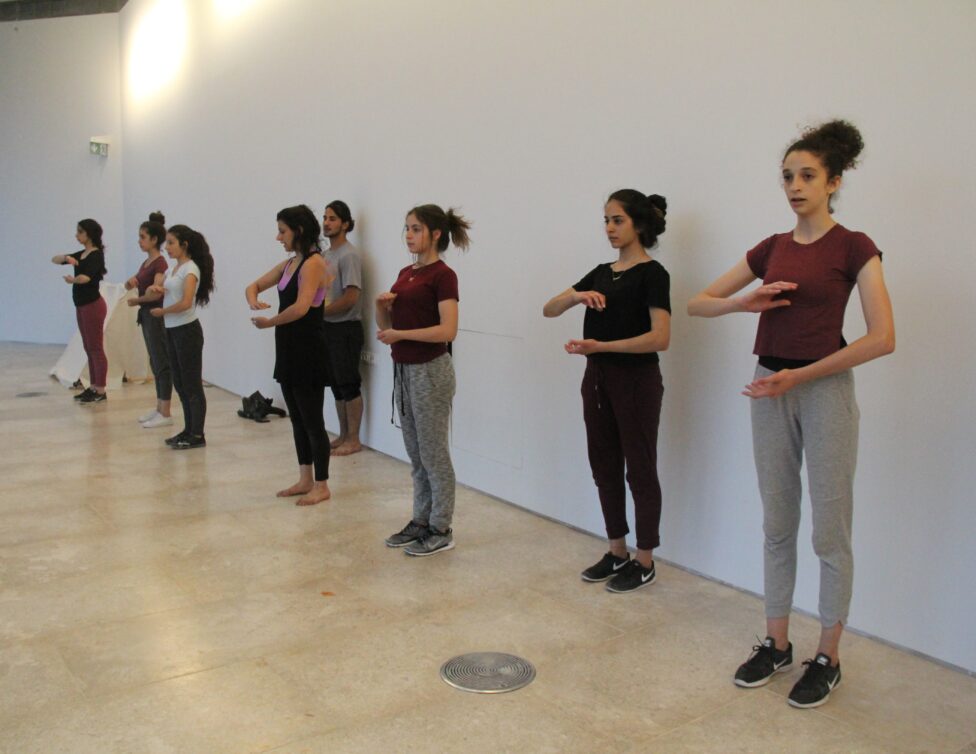In a dance studio with wooden floors and white walls, several dancers in a line face the same direction with arms folded close to the body.