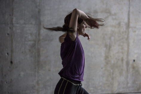 Photo of Jimena Paz in movement. Her arms are raised with her elbows bent. She wears a purple t-shirt and dark pants. Her hair whips around her face. Photo by Maria Baranova.