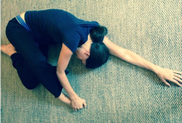 Photo of Jimena Paz lying on the floor in a fetal position with one arm extended above her head. She has dark hair and wears dark colored clothing. Photo courtesy of the artist.