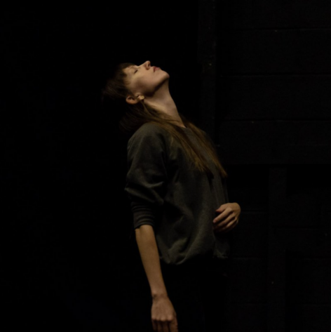 Standing before a shadowy background, Katie wears dark-colored clothing and tips her head backward with eyes closed.