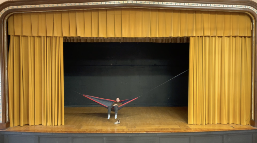 A figure lays in a hammock suspended across a stage, framed by yellow curtains