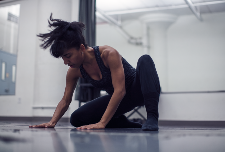 A photo of Megan Curet dancing in a studio. She is in a crouched position using her arms for support and looking down. She wears a black tank top and leggings. Photo by Edward Pages.